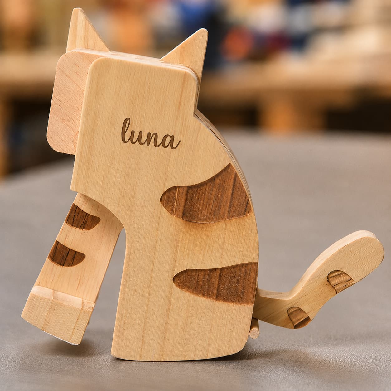 A wooden cat-shaped object, likely a toy, book, or decorative item, engraved with the name 'luna' on its side. The cat is crafted from light-colored wood with darker brown inlay patterns, set against a blurred background.