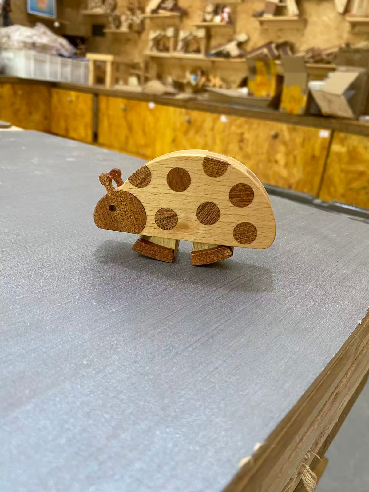 A small wooden ladybug toy with a light natural wood body and darker brown circular spots, standing on a gray tabletop in a workshop with wooden shelves and tools in the background.
