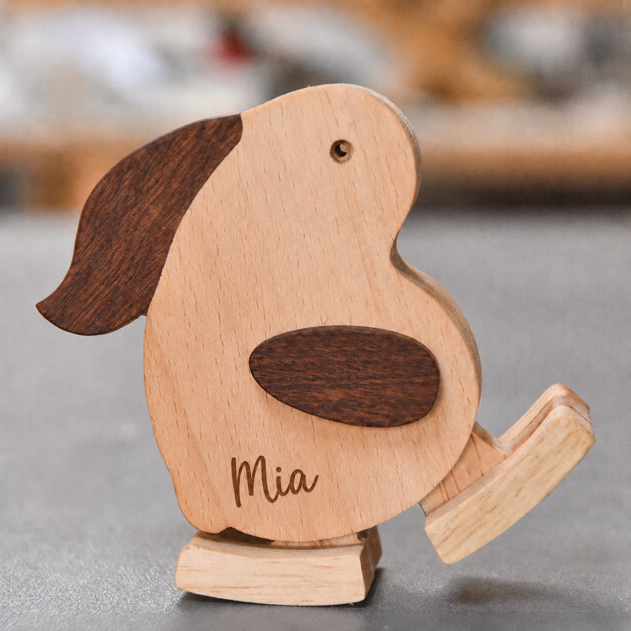 A personalized wooden rocking dog toy with a light natural wood body, dark brown ears and wing detail, and the name 'Mia' engraved on the front, resting on a gray surface.