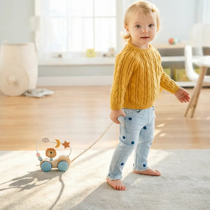 Child in a yellow sweater and blue polka dot pants standing on a rug with a toy in a room.
