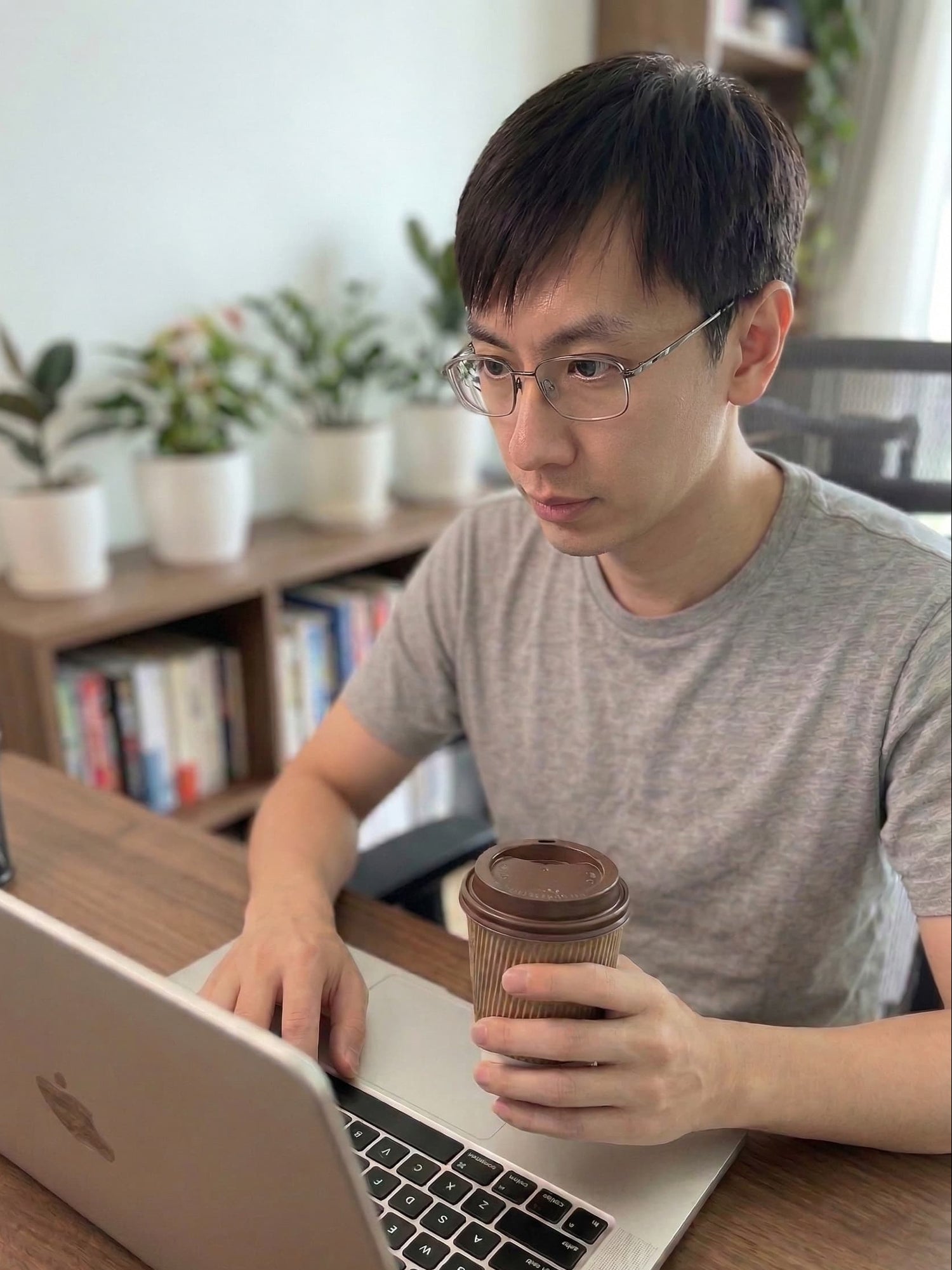 Man sitting at a desk with a laptop and holding a coffee cup, surrounded by plants and books.
