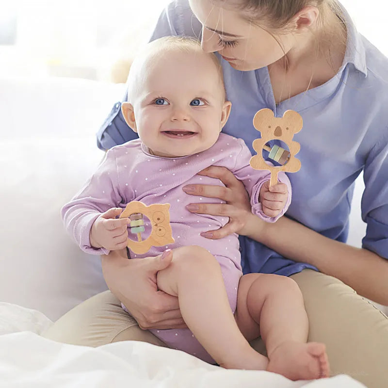 Baby holding a wooden teething toy with a woman sitting next to them on a white couch.