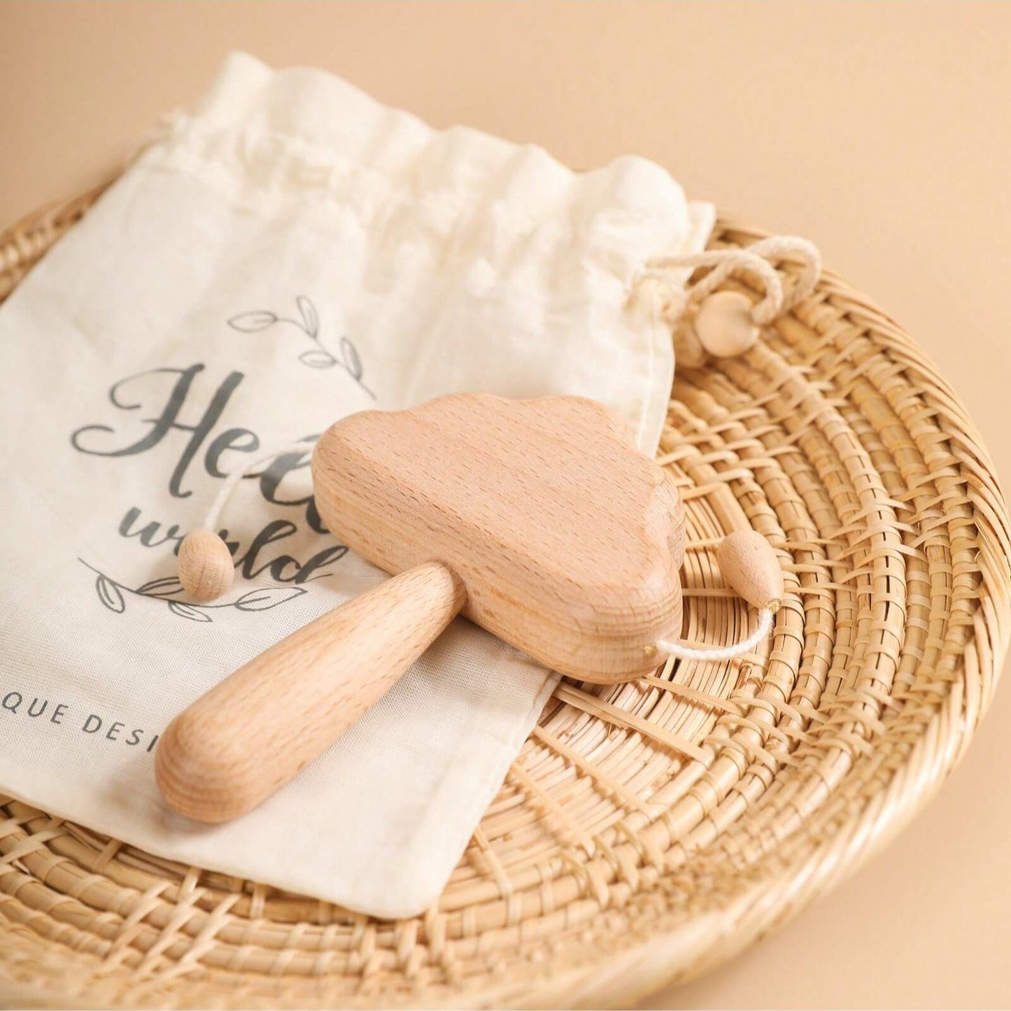 Wooden baby rattle on a white cloth with a drawstring bag labeled 'Hello world' on a woven mat.