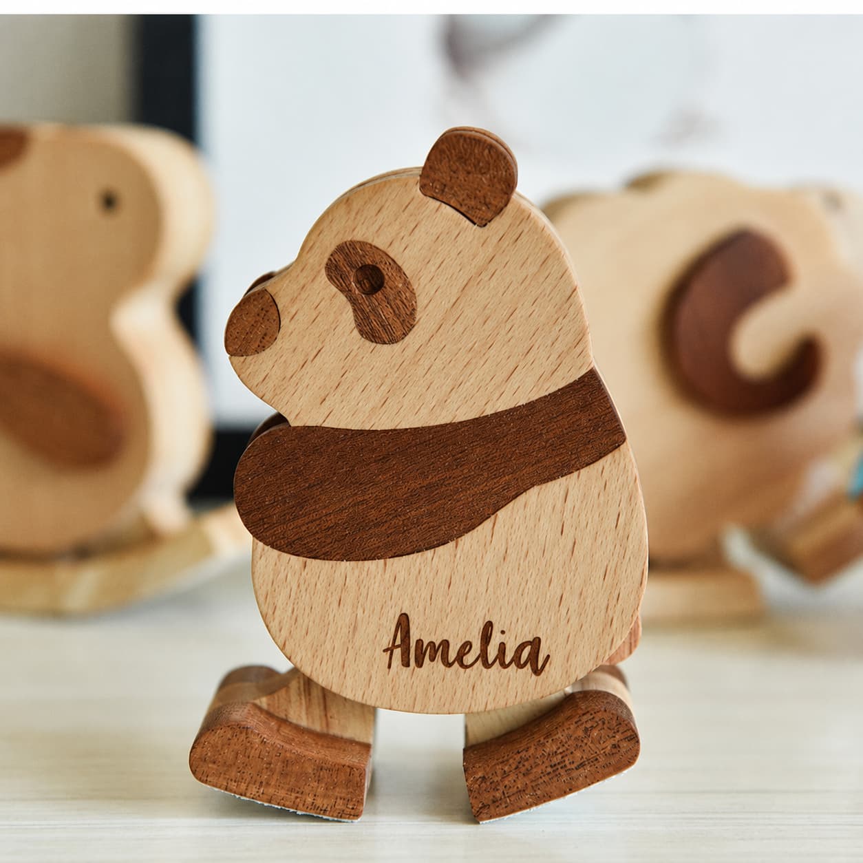 A personalized wooden panda toy with the name 'Amelia' engraved on its belly, standing on a light surface with other blurred wooden animal toys in the background.