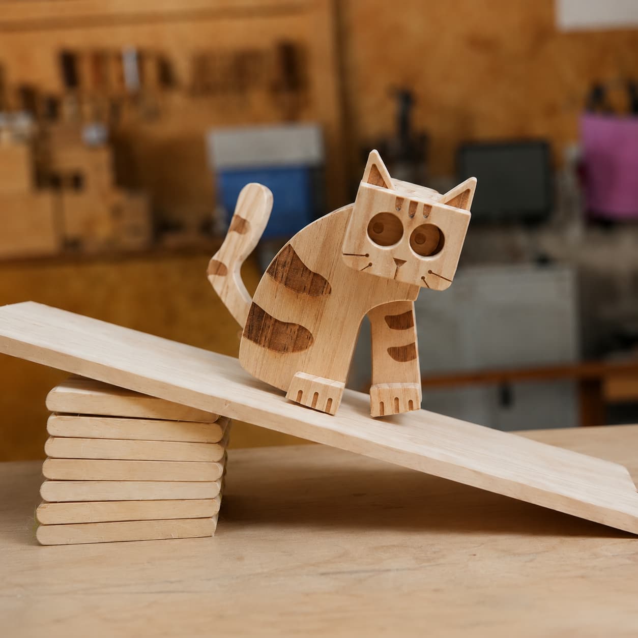 Handmade wooden cat ramp walker toy with dark walnut striped accents and large hollow eyes, walking down a mechanical gravity-powered ramp in a carpentry workshop.