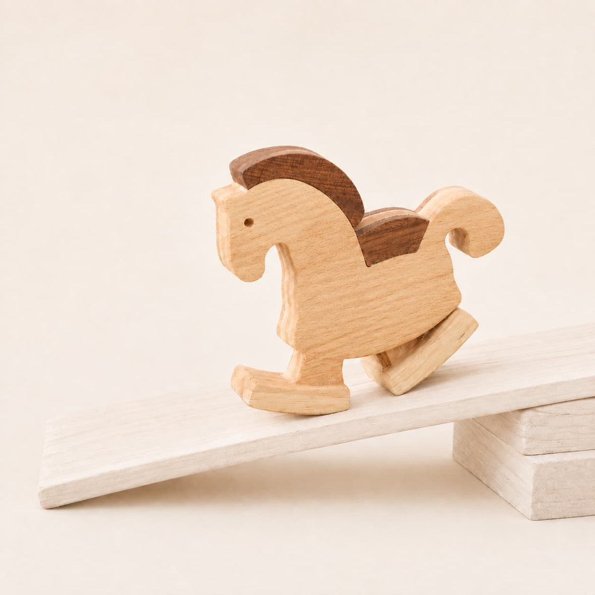 A minimalist wooden rocking horse ramp walking toy with a natural light wood body and a dark brown mane, shown balancing on a small wooden ramp against a warm cream background.