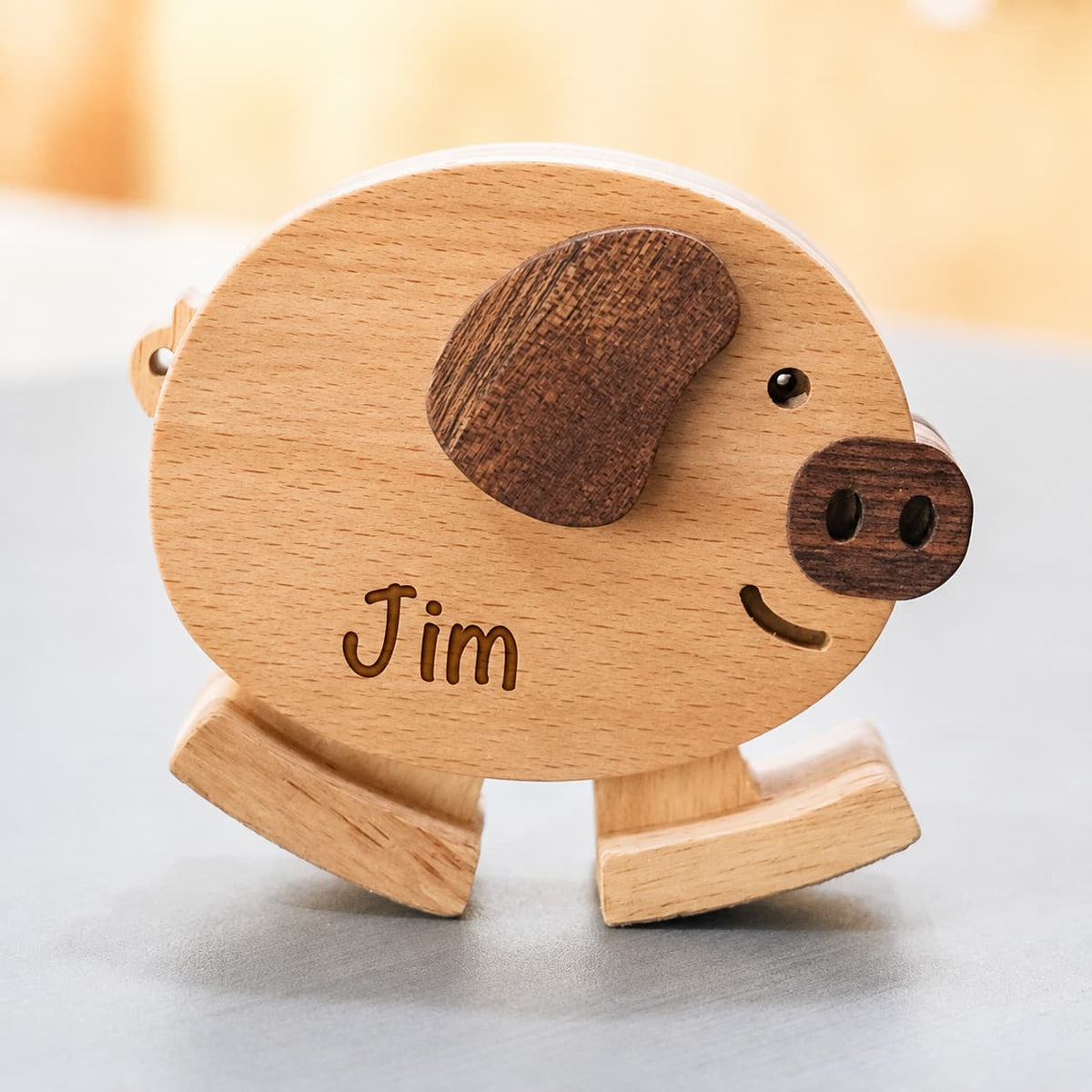 A personalized wooden pig toy with a light natural wood body, dark brown ears and snout, and the name 'Jim' engraved on the front, shown on a neutral gray surface with a warm, blurred background.
