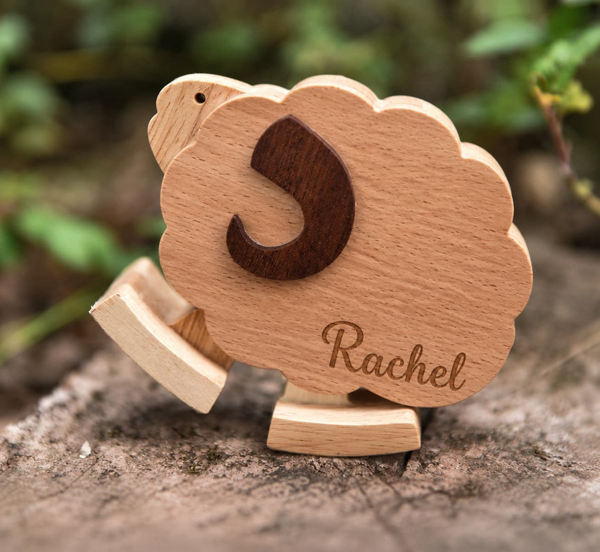 A personalized wooden sheep toy with the name 'Rachel' engraved on its body and a dark wooden letter 'S' on its back, displayed on a tree trunk with a blurred natural background.