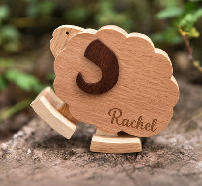 A personalized wooden sheep toy with the name 'Rachel' engraved on its body and a dark wooden letter 'S' on its back, displayed on a tree trunk with a blurred natural background.