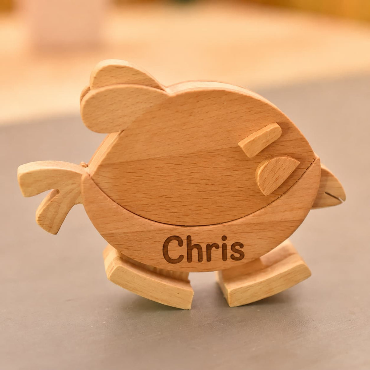 A personalized wooden toy bird with the name 'Chris' engraved on its side, resting on a neutral gray surface with a warm, softly blurred background.