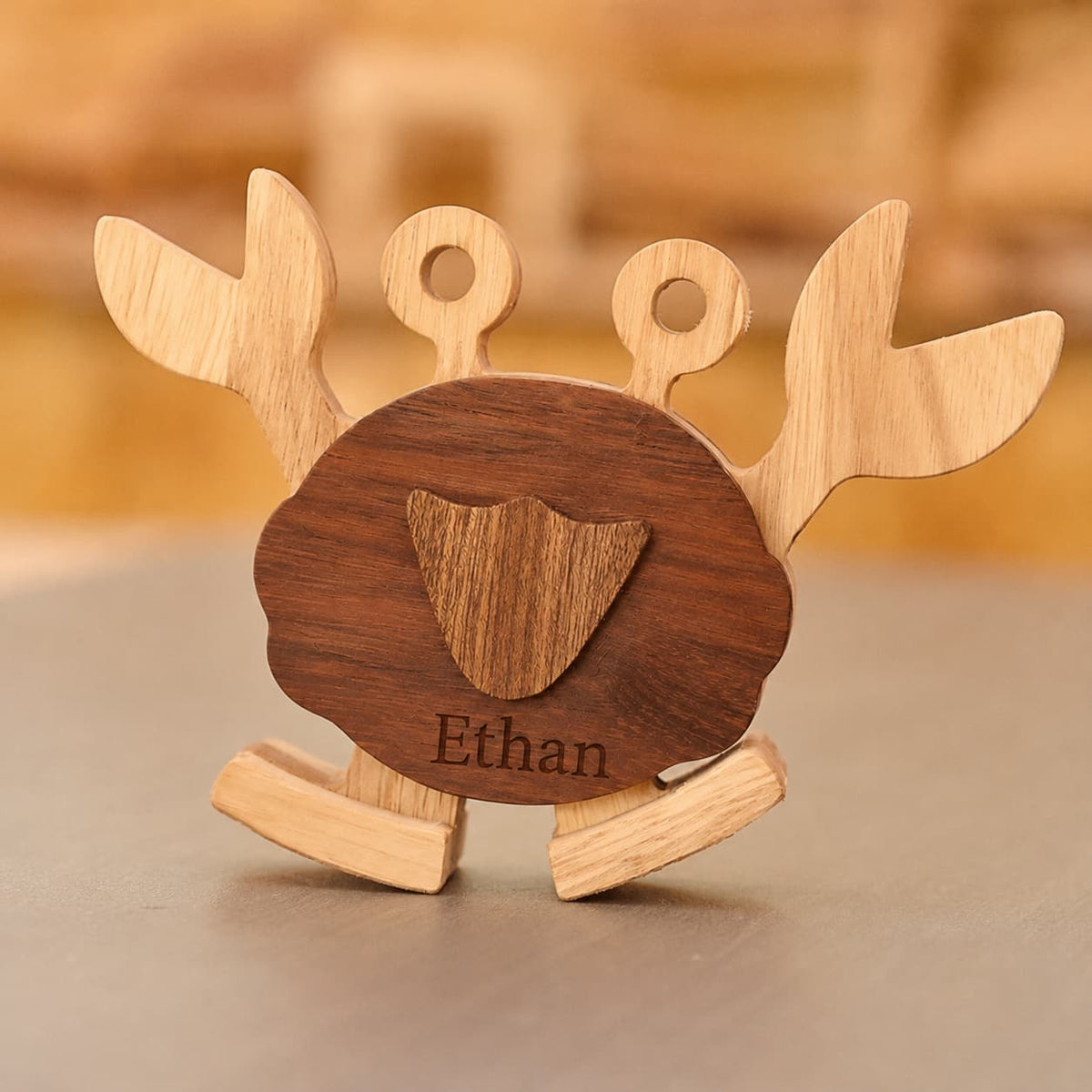 A personalized wooden toy crab with a dark brown engraved shell and light natural wood legs, antennae, and claws, standing on a gray surface with a warm, blurred background.