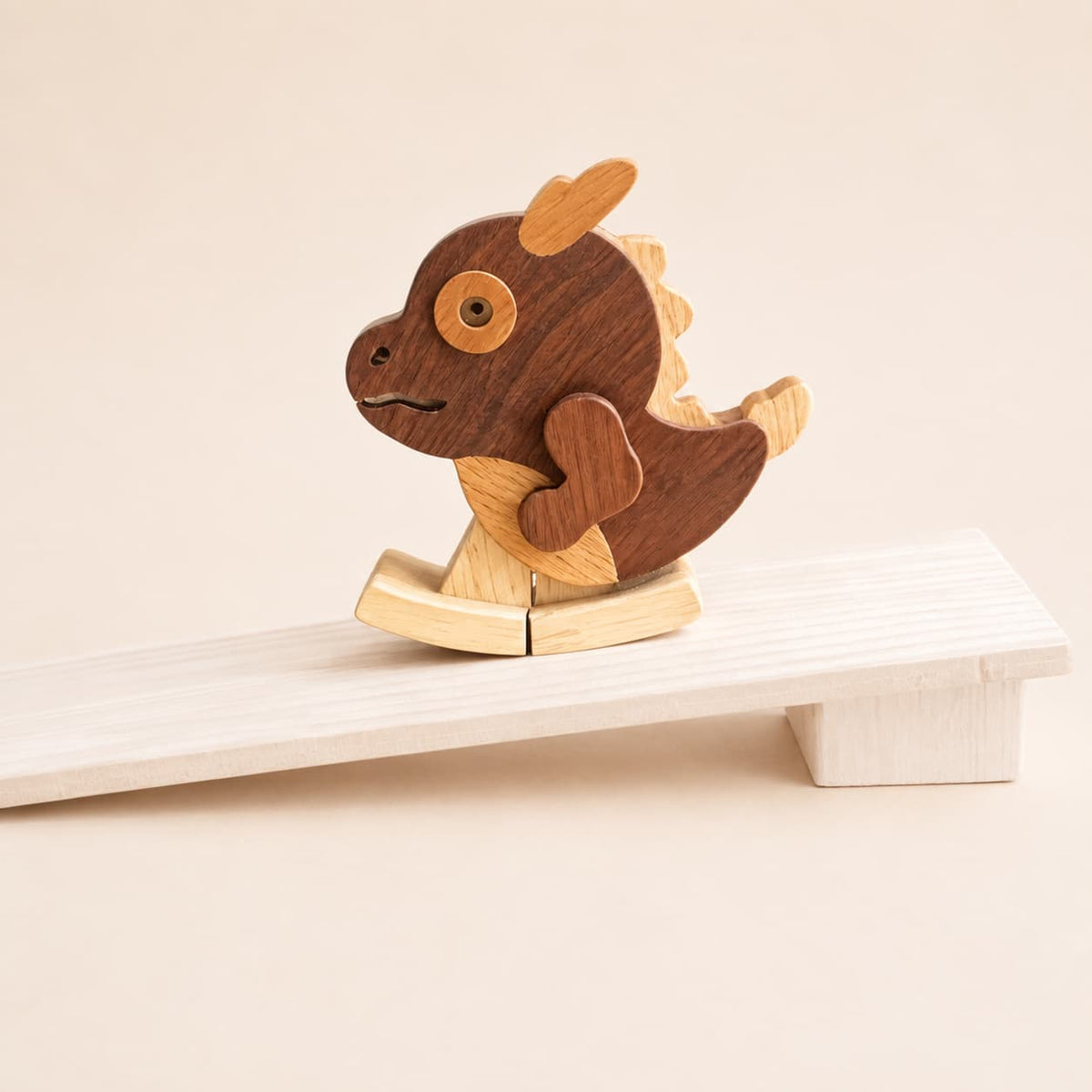 A wooden rocking dragon toy with a dark brown body, light tan fins and belly, resting on a white wooden rocking board against a warm beige background.