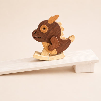 A wooden rocking dragon toy with a dark brown body, light tan fins and belly, resting on a white wooden rocking board against a warm beige background.