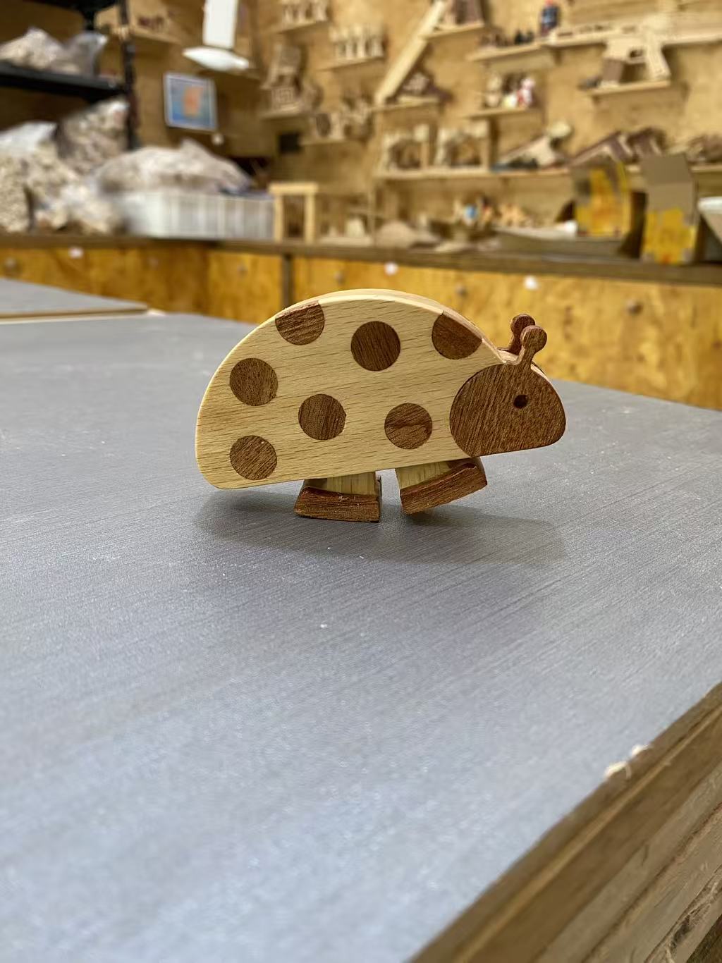 A small wooden ladybug toy with natural wood spots, standing on a gray tabletop inside what appears to be a wooden craft or toy workshop.