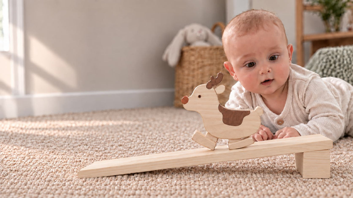 A baby lying on a beige carpet in a nursery setting, interacting with a wooden toy balance board featuring a small rocking reindeer.