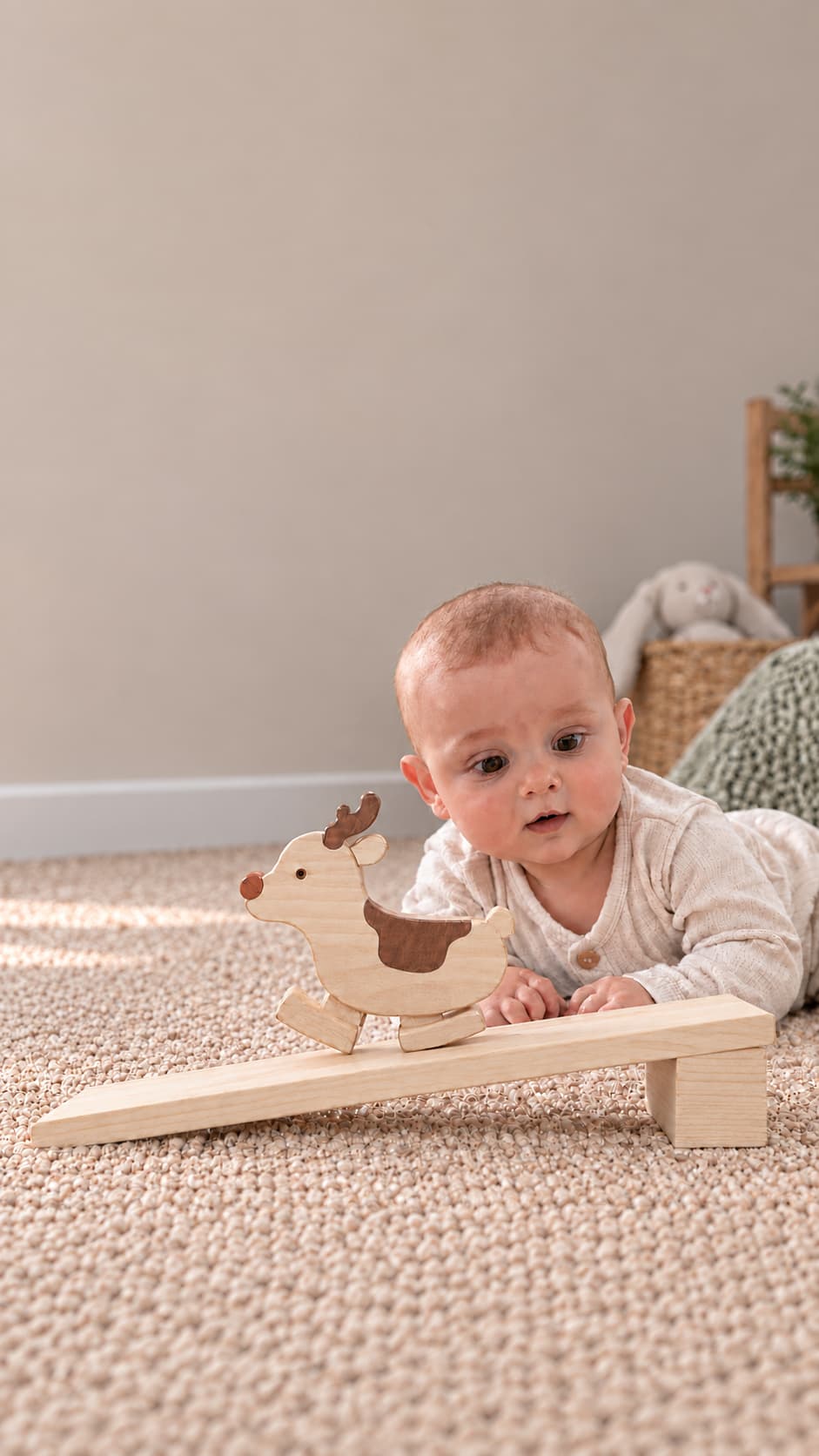 A baby lying on a beige carpet plays with a wooden toy balance board featuring a reindeer-shaped weight, in a softly lit nursery setting.