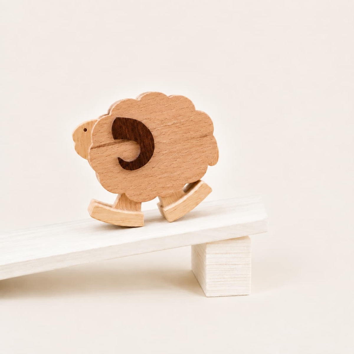 A small wooden sheep ramp walking toy with a dark brown cutout face, shown balancing on a white wooden seesaw against a warm off-white background.